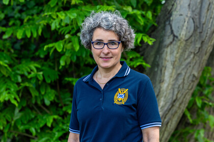 Susanne Quincke steht mit blauem Polo-Shirt vor einer grünen Blätterwand und lächelt in die Kamera.