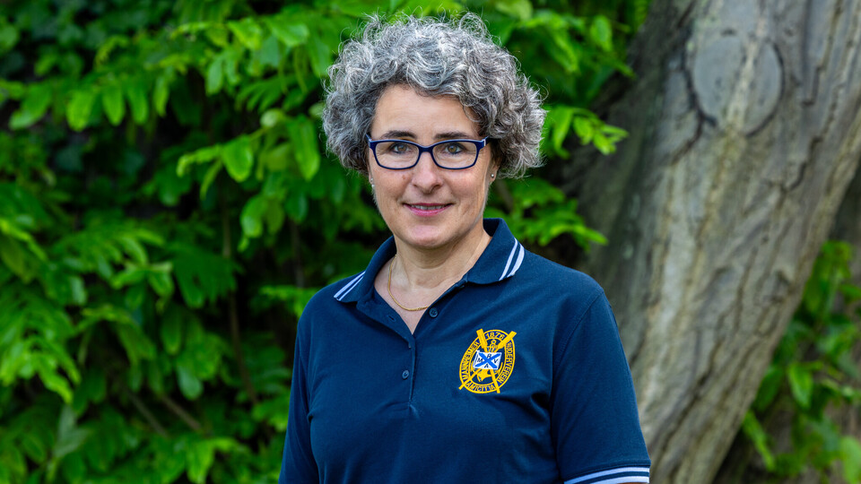 Susanne Quincke steht mit blauem Polo-Shirt vor einer grünen Blätterwand und lächelt in die Kamera.