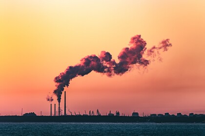 Industrielandschaft am Wasser mit Schornsteinen, aus denen Rauch unter einem orangefarbenen Himmel aufsteigt.