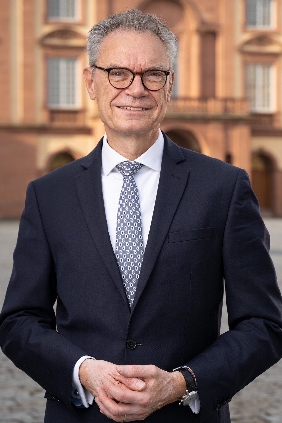 Joachim Lutz hat kurze Haare und eine Brille. Er trägt ein weißes Hemd, eine Krawatte und einen dunklen Anzug und steht vor dem Schloss.