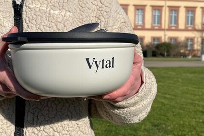 Die hellgraue Mehrweg-Bowl von Vytal mit schwarzem Deckel wird hochgehalten