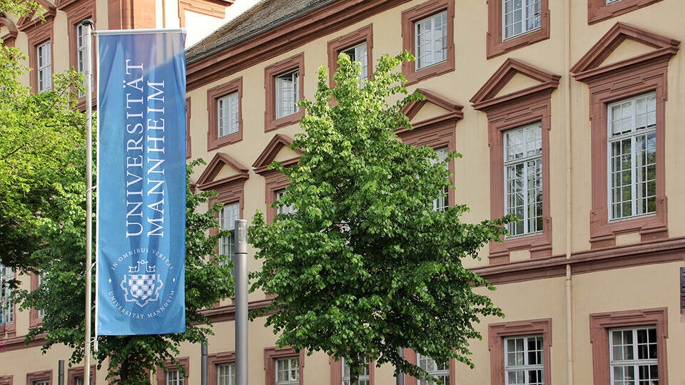 Eine blaue Fahne mit dem Logo der Uni Mannheim weht vor dem Haupteingang der Universität im Ostflügel des Schlosses.