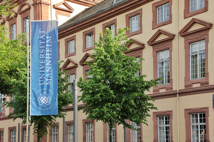Eine blaue Fahne mit dem Logo der Uni Mannheim weht vor dem Haupteingang der Universität im Ostflügel des Schlosses.