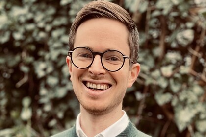 Florian Pethig. He is wearing a green sweater and is standing in front of a green hedge.