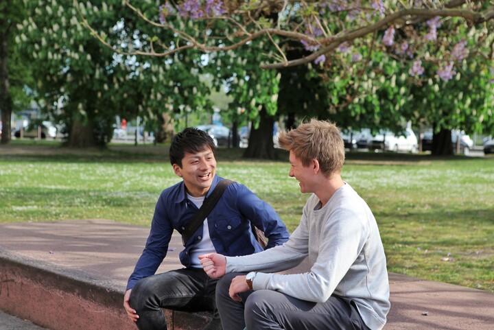 Zwei Studenten sitzen auf Steinen und lachen gemeinsam. Im Hintergrund blühen ein paar Bäume.