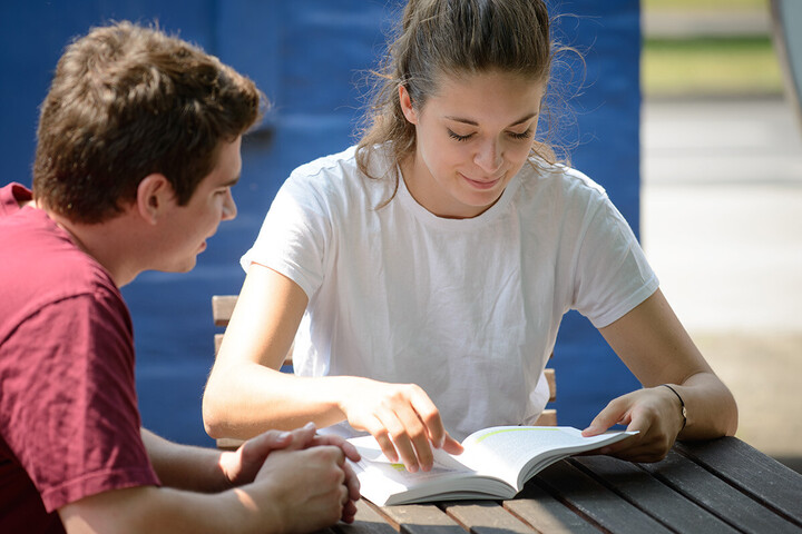 Zwei Studenten sitzen im Außenbereich des Café Soleil und lesen gemeinsam in einem Buch.