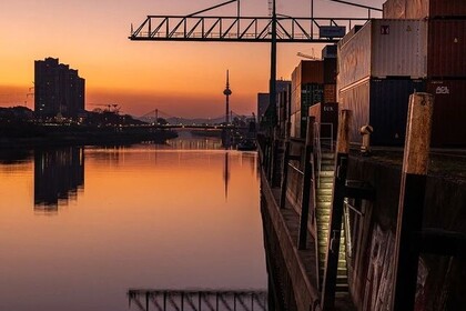 Sonnenuntergang über dem Neckar, am rechten Ufer Container und ein Kran im Hafenviertel. Im Hintergrund die Silhouette der Stadt mit Fernmeldeturm, Hochäusern und Collini-Steg.