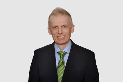 Prof. Maug hat kurze Haare und trägt ein dunkles Jackett, ein hellblaues Hemd und eine grüne Krawatte