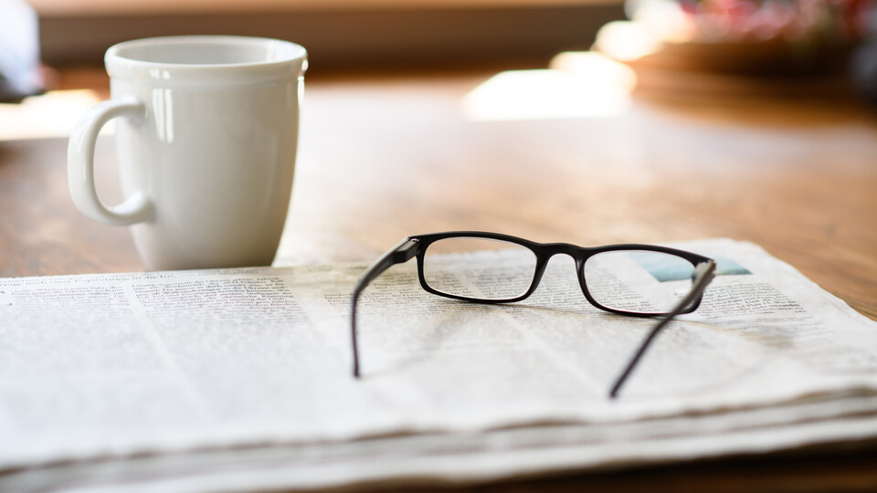 Auf einem Tisch liegt eine Zeitung und eine Brille neben einer Kaffeetasse