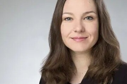 Portrait von Tanja Skambraks. Sie trägt eine schwarze Bluse und einen dunkelbraunen Blazer und steht vor einer grauen Wand.