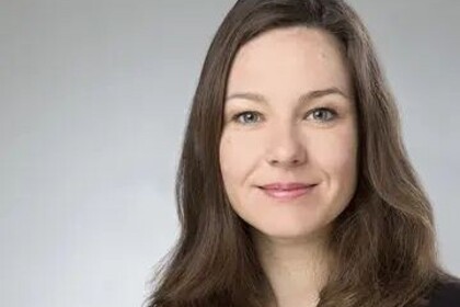 Portrait von Tanja Skambraks. Sie trägt eine schwarze Bluse und einen dunkelbraunen Blazer und steht vor einer grauen Wand.
