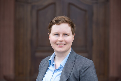 Portrait of Theresa Buehrle. She is wearing a light blue shirt with a gray blazer over it and is standing in front of Mannheim Palace.