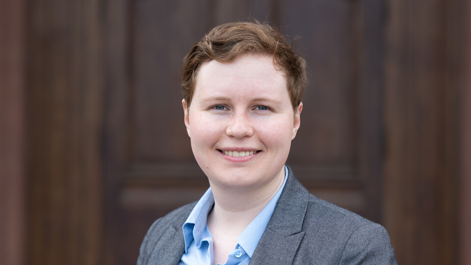 Portrait of Theresa Buehrle. She is wearing a light blue shirt with a gray blazer over it and is standing in front of Mannheim Palace.