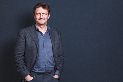 Portrait von Peter Vorderer. Er trägt ein blaues Hemd sowie einen grauen Blazer und steht vor einer dunkelblauen Wand.