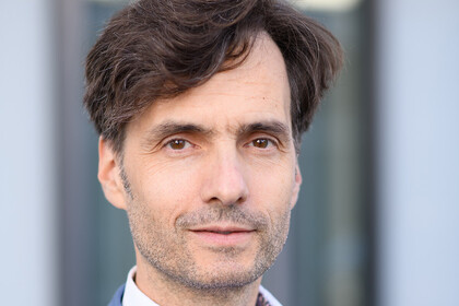 Georg Alpers hat kurze braune Haare, einen Dreitagebart und trägt einen blauen Anzug mit weißem Hemd.