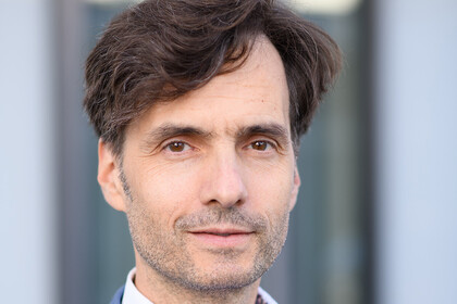 Georg Alpers hat kurze braune Haare, einen Dreitagebart und trägt einen blauen Anzug mit weißem Hemd.