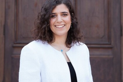 Amina Elbarbary hat mittellange, lockige und braune Haare. Sie trägt ein dunkles Oberteil und einen weißen Blazer darüber.