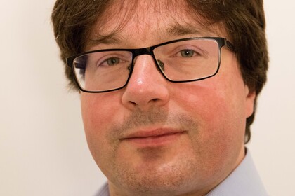 Portrait von Philipp Fischinger. Er trägt ein hellblaues Hemd und steht lächelnd vor einer weißen Wand.