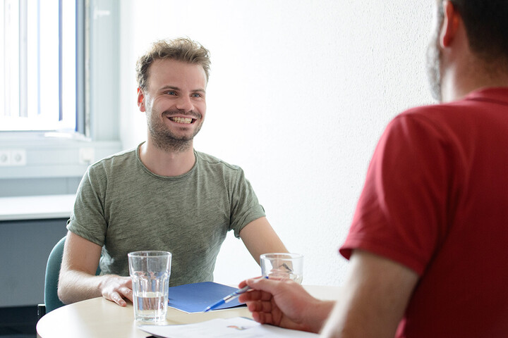 Smiling man, dressed in a green T-shirt, sits at a round table opposite another person, dressed in a red T-shirt, who is holding a ballpoint pen in his right hand.  There are two glasses of water and folders on the table. 