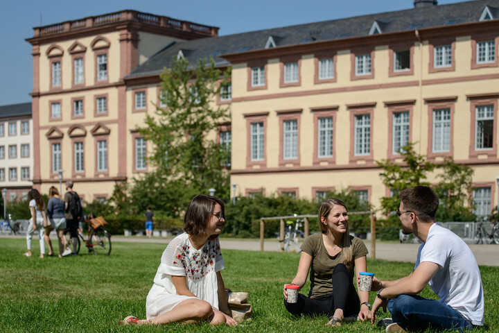 Zwei Studentinnen und ein Student sitzen auf einer grünen Wiese. Sie tragen frühlingshafte Kleidung und halten Kaffee-To-Go-Becher in ihren Händen.