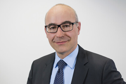 Portrait von Achim Wambach. Er trägt ein hellblaues Hemd sowie eine dunkelblau karrierte Krawatte und einen schwarzen Blazer. Er steht lächelnd vor einer grauen Wand.