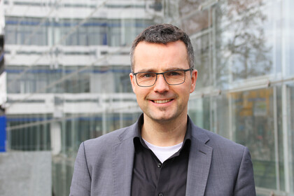 Portrait von Marc Debus. Er trägt ein schwarzes Hemd sowie einen grauen Blazer und steht vor dem A5-Gebäude.
