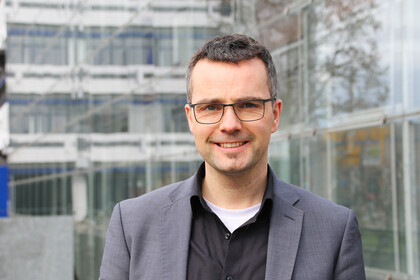 Portrait von Marc Debus. Er trägt ein schwarzes Hemd sowie einen grauen Blazer und steht vor dem A5-Gebäude.