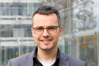 Portrait von Marc Debus. Er trägt ein schwarzes Hemd sowie einen grauen Blazer und steht vor dem A5-Gebäude.