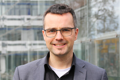 Portrait von Marc Debus. Er trägt ein schwarzes Hemd sowie einen grauen Blazer und steht vor dem A5-Gebäude.