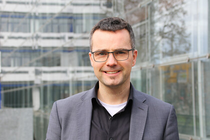Marc Debus trägt ein schwarzes Hemd mit einem grauen Sacko und eine Brille. Er steht vor dem Unigebäude in A5.