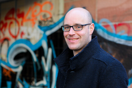 Portrait von Leif Döring. Er trägt einen dunkelblauen Mantel und steht vor einer Graffiti-Wand.