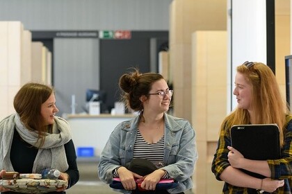 Drei Studierende unterhalten sich und halten Uni-Utensilien wie Laptops und Bücher in den Händen