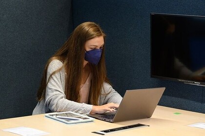 Eine Studentin sitzt in der Bibliothek vor ihrem Laptop. Sie hat lange braune Haare und trägt eine blaue FFP2-Maske.