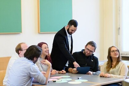 Aufnahme einer Gruppe von Personen, die zusammen an einem Tisch sitzen und sich Notizen machen. Eine Person steht dazwischen und zeigt auf etwas.