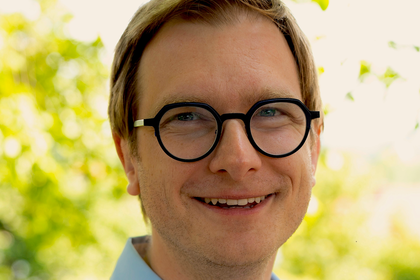 Portrait of Benjamin Engst. He is wearing a light blue shirt and standing in front of a tree.