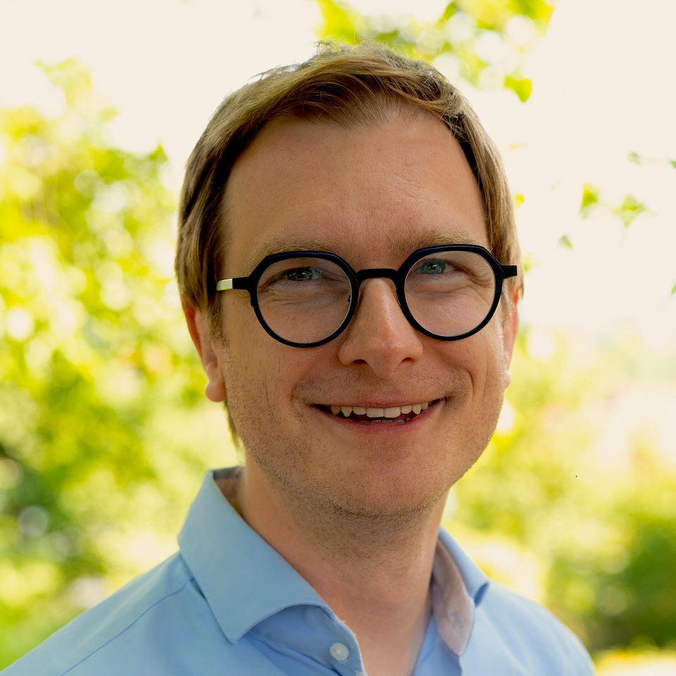 Portrait of Benjamin Engst. He is wearing a light blue shirt and standing in front of a tree.