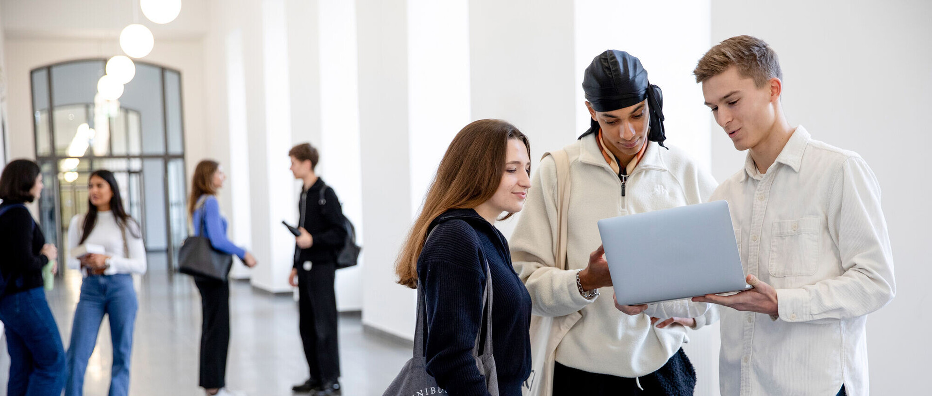 A group of students is chatting in the hallway of the castle building. One female student and two male students are looking at something on a laptop.