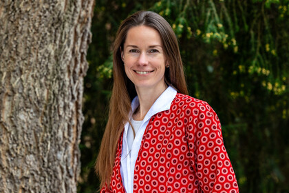 Prof. Dr. Ilze Plavgo. Sie steht lächelnd vor einem Baum und trägt einen rot gemusterten Blazer.