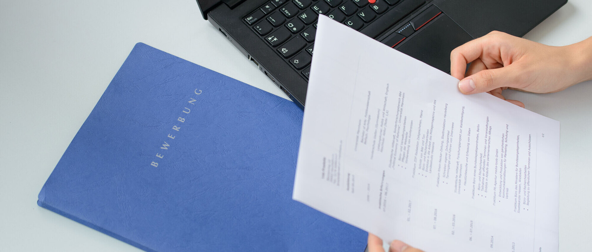 A job application is being held above a blue application folder and a black laptop