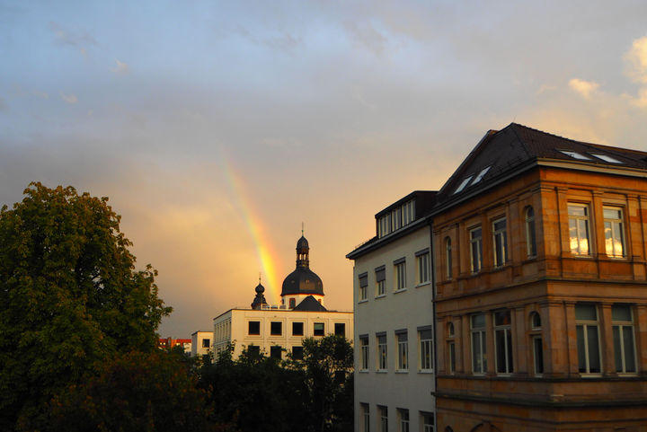 In goldenem Licht steht die Sternenwarte von Mannheim. Im Vordergrund sind Wohnhäuser, im Hintergrund Wolken und ein Regenbogen.