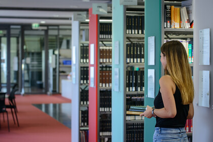 Studierende mit langen blonden Haaren läuft durch die Bibliothek und trägt Bücher in den Händen.