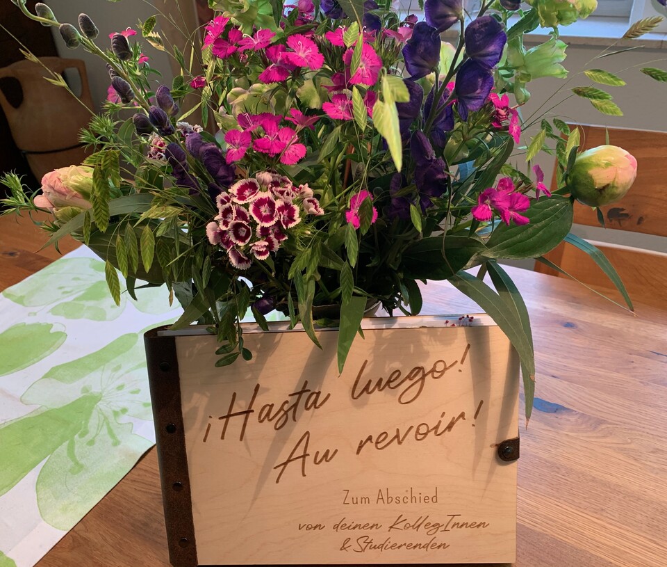 Im Hintergrund steht eine Vase mit Blumen. Vor der Vase aufgestellt steht ein Buch mit hölzernem Einband, mit der Aufschrift "Hasta luego!, "Au revoir".
