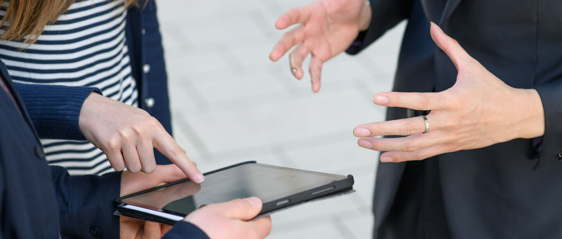 Eine Person hält ein Tablet in den Händen. Eine zweite Personen zeigt auf den schwarzen Bildschirm des Tablets. Eine dritte Persob gestikuliert mit den Händen in der Luft.