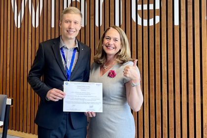 Marc wears a dark suit, is holding an award certificate and is standing next to Professor Ruth V. Aguilera who is wearing a light grey dress.