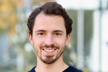 Philipp Kadel hat wellige dunkelbraune Haare und einen Bart. Er steht im Freien vor einem Gebäude und einem Baum und trägt einen dunkelblauen Pullover.