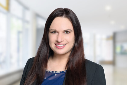 Portrait von Anne Hannusch. Sie trägt eine blaue Bluse sowie einen schwarzen Blazer und steht lächelnd vor einer Fensterfront.