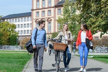 Drei Studierende laufen auf der Mensawiese. Die Person links hat kurze braune Haare und trägt ein blaues Oberteil. Die Person in der Mitte schiebt ihr Fahrrad neben sich her und hat lange blonde Haare. Sie trägt eine beige Jacke. Die Person rechts hat lange braune Haare und trägt ein weißes Oberteil mit einer roten Jacke.