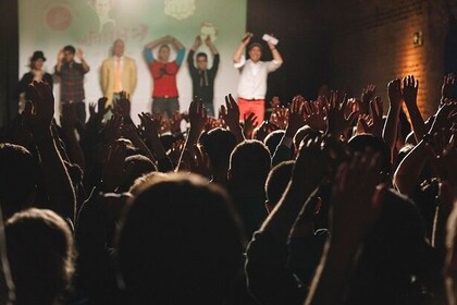 Im Hintergrund stehen Personen auf der Bühne. Im Vordergrund sieht man ein Publikum. Alle Menschen halten ihre Hände in die Lüfte. 