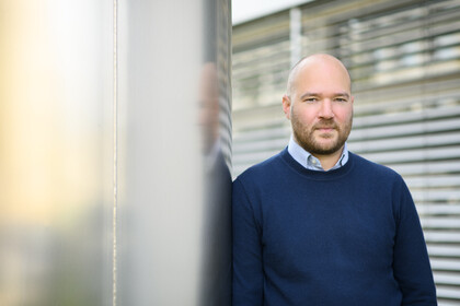 Eine Person trägt ein weiß-blau gestreiftes Hemd unter einem dunkelblauen Pullover und steht angelehnt an eine Säule. Die Person heißt Richard Traunmüller.