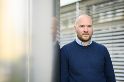 Eine Person trägt ein weiß-blau gestreiftes Hemd unter einem dunkelblauen Pullover und steht angelehnt an eine Säule. Die Person heißt Richard Traunmüller.